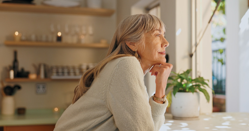 Older woman resting her elbow on her kitchen counter, gazing out of the window with a smile on her face.