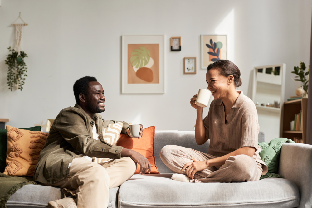 A young couple at home drinking coffee on their sofa, in conversation.