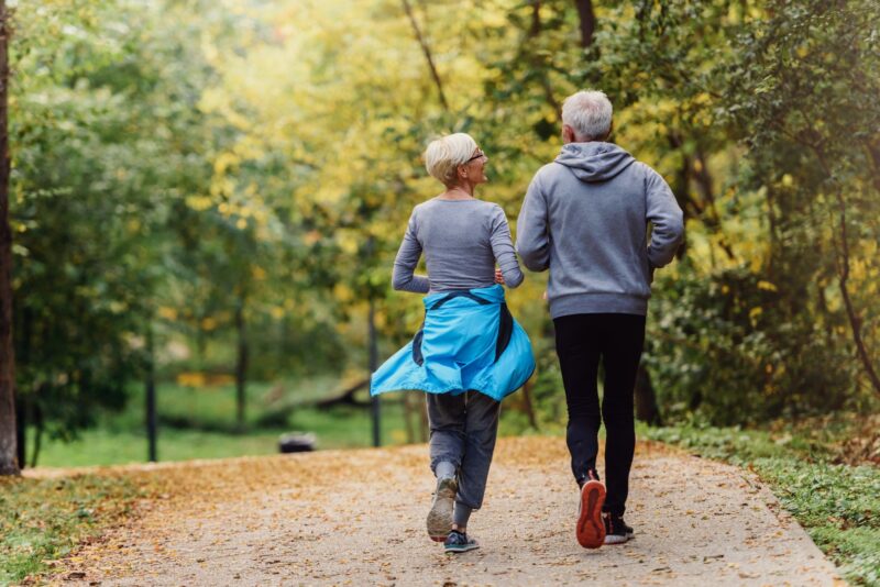 elderly couple, woman and man, wearing grey athletic wear running in woods.
