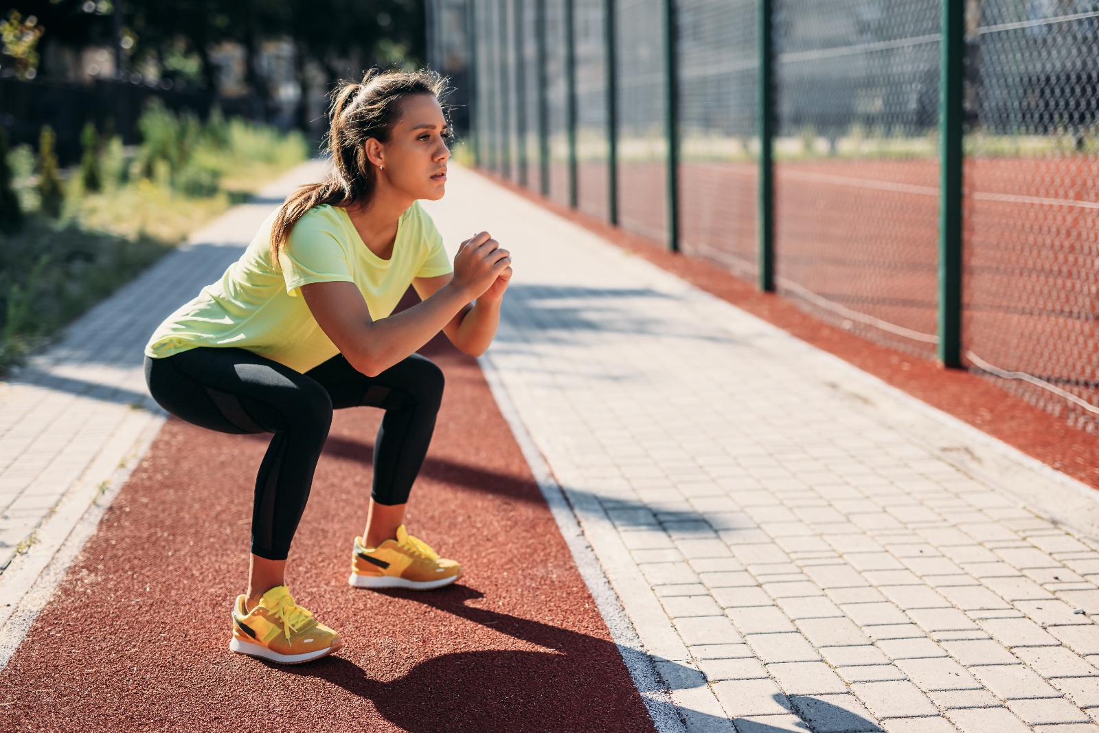 Brunette woman with hair in a pony tail wearing yellow top and trainers performing a squat at a running track