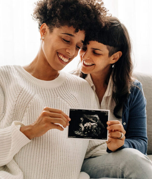 Same-sex pregnant couple holding up their ultrasound scan. Expectant lesbian couple smiling cheerfully while holding an ultrasound picture of their unborn baby. Young queer couple expecting a baby.