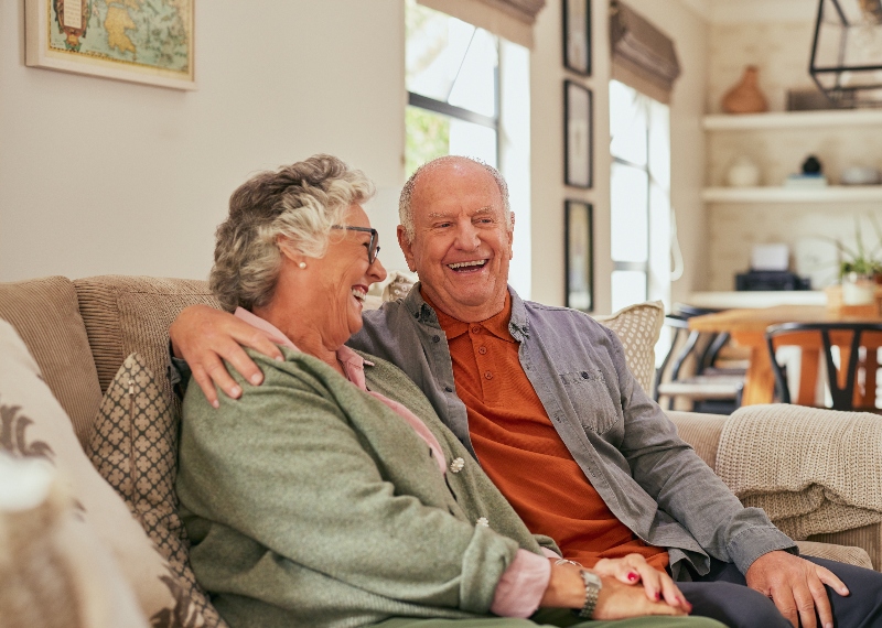 Smiling senior couple sitting on couch while having a pleasure conversation at home. Elder man and old beautiful woman sharing an happy moment sitting on a cozy sofa. Relaxed elderly couple enjoying a warm and cheerful discussion in their living room.