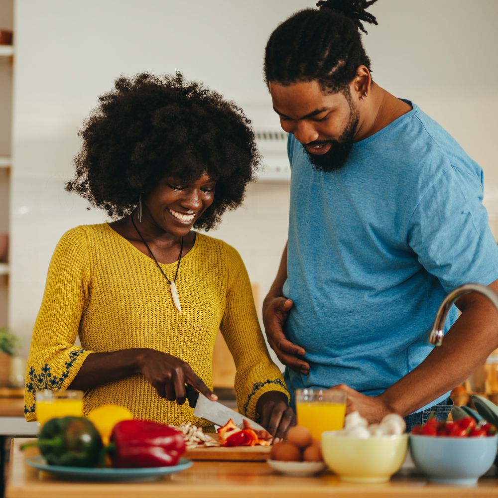 A cheerful interracial woman is chopping and cooking vegetables in a kitchen at home while her hungry husband is stealing food and trying out it. A married couple in the kitchen at home together.