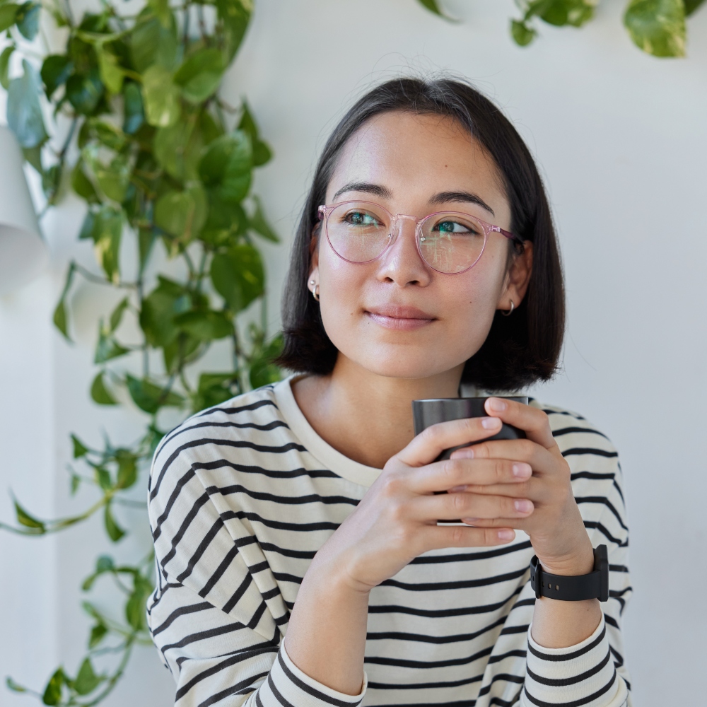 Vertical shot of thoughtful young Asian woman wears transparent eyeglasses and striped jumper drinks coffee looks away sits at white desk with modern technologies around against cozy interior.